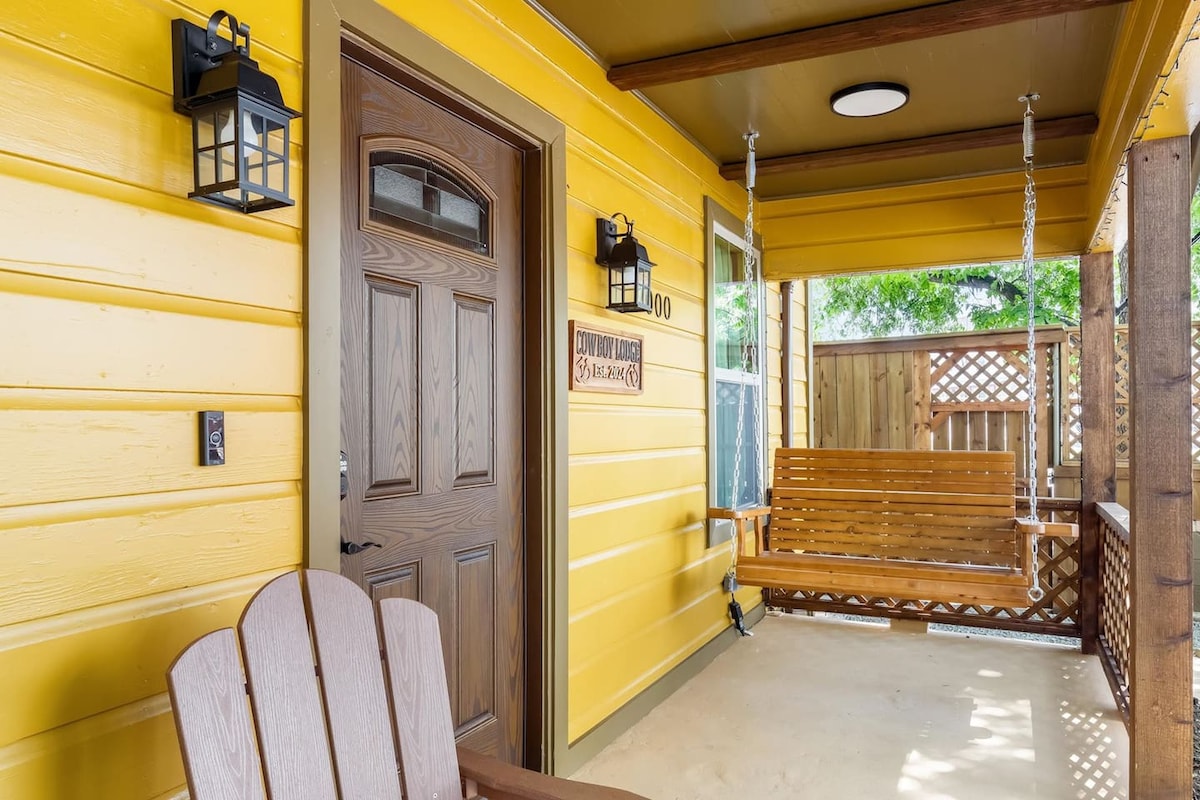 A welcoming front porch features a bright yellow facade, accented by a wooden door and outdoor lanterns. A swing chair provides a cozy spot for relaxation, while nearby windows allow natural light to fill the space, enhancing the inviting atmosphere of the entrance.