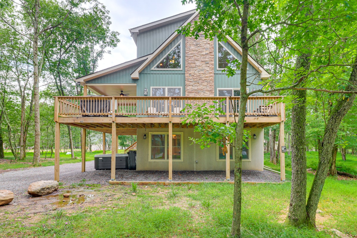 The exterior of the cabin features a wraparound deck with a wooden railing, surrounded by lush greenery. Large windows allow natural light to enter the home, and a hot tub is visible on the deck, nestled in a serene forest setting.