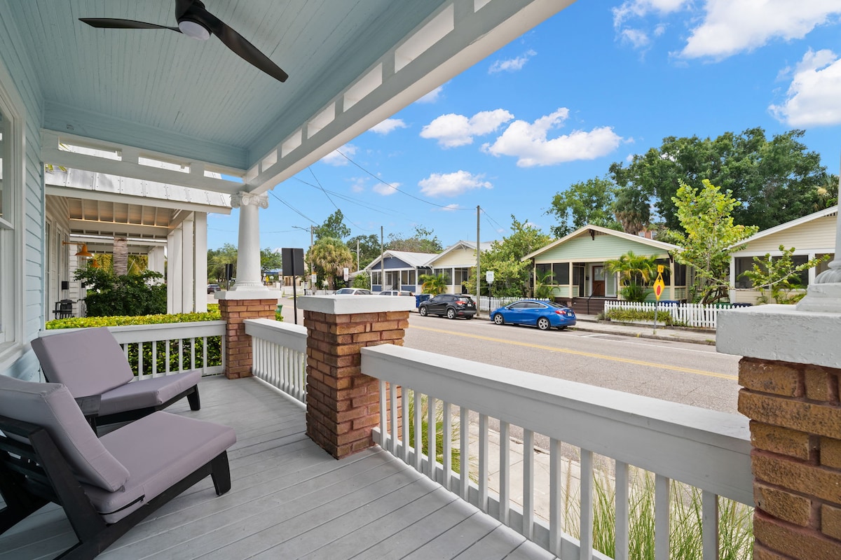 A spacious front porch features two lounge chairs facing the street, offering a comfortable view of the neighborhood. The porch is framed by charming brick columns, and a light blue ceiling adds a touch of character. Lush greenery and other homes can be seen in the surroundings.