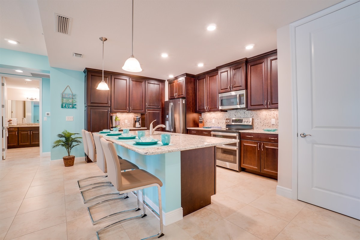 A modern kitchen features dark wooden cabinetry contrasted by light granite countertops. Bar seating with light-colored stools stretches along one side. Stainless steel appliances, including a refrigerator and oven, are visible, with subtle under-cabinet lighting enhancing the ambiance.