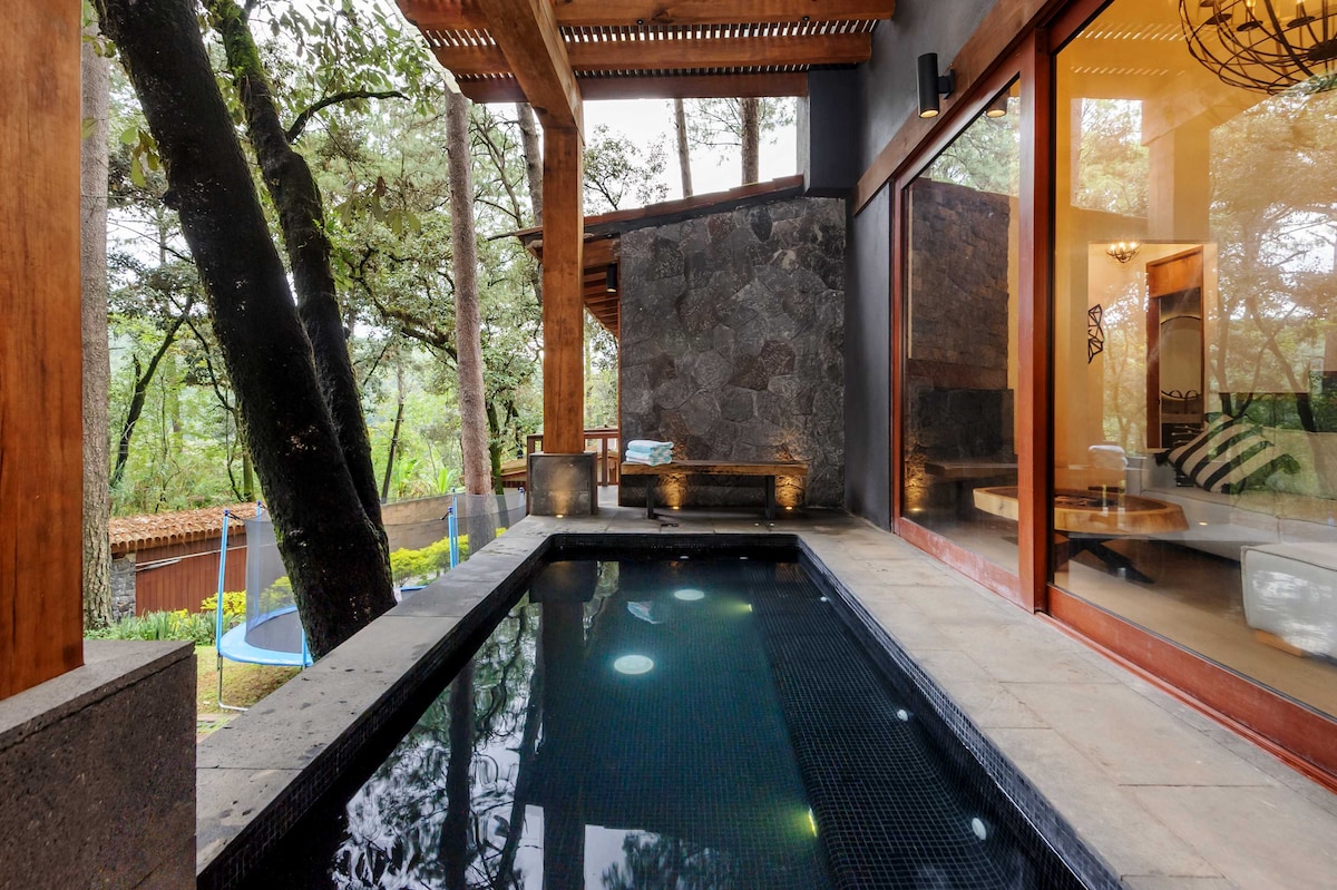 A private outdoor Jacuzzi is set against a backdrop of trees, surrounded by natural stone walls and wood accents. Large glass panels provide views of the serene landscape, with soft lighting reflecting off the water's surface, creating a tranquil atmosphere.