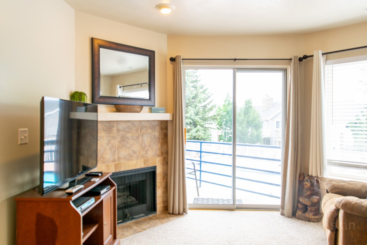 A comfortable living area features a flat-screen television positioned on a wooden stand. A modern fireplace is integrated into the corner, and natural light enters through sliding glass doors that lead to a balcony. Soft furnishings add to the cozy atmosphere.