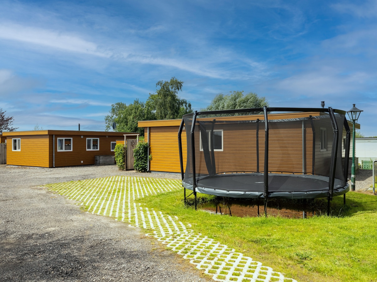Two chalets featuring wooden exteriors are viewed from a serene outdoor space. A round trampoline is situated on a grassy area, with a pathway of white bricks leading through the grounds. The backdrop includes trees and a clear blue sky, enhancing the peaceful environment.