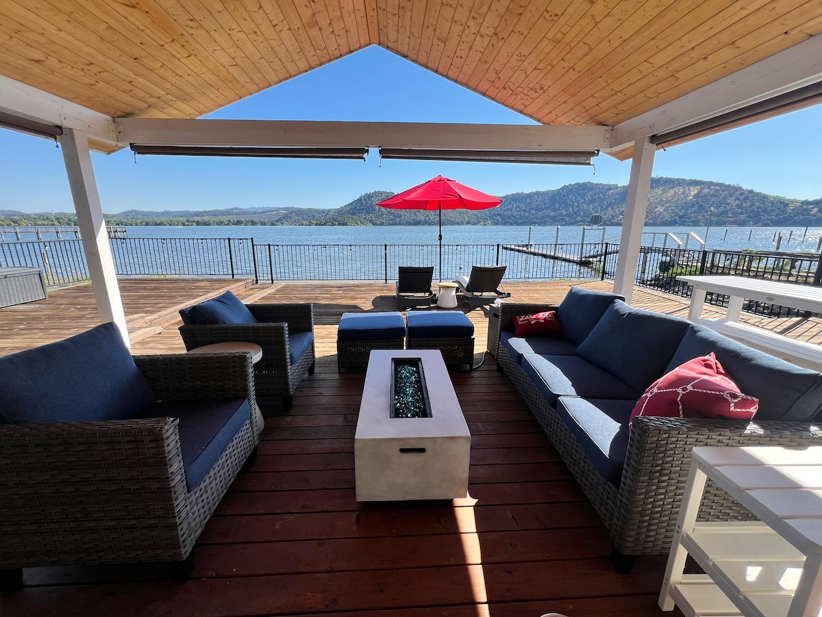 An outdoor deck features comfortable seating arrangements, including two armchairs and a spacious sofa with accent pillows. A central fire table adds ambiance, while an umbrella provides shade. The scenic lakeshore view is framed by rolling hills in the background, enhancing the tranquil setting.