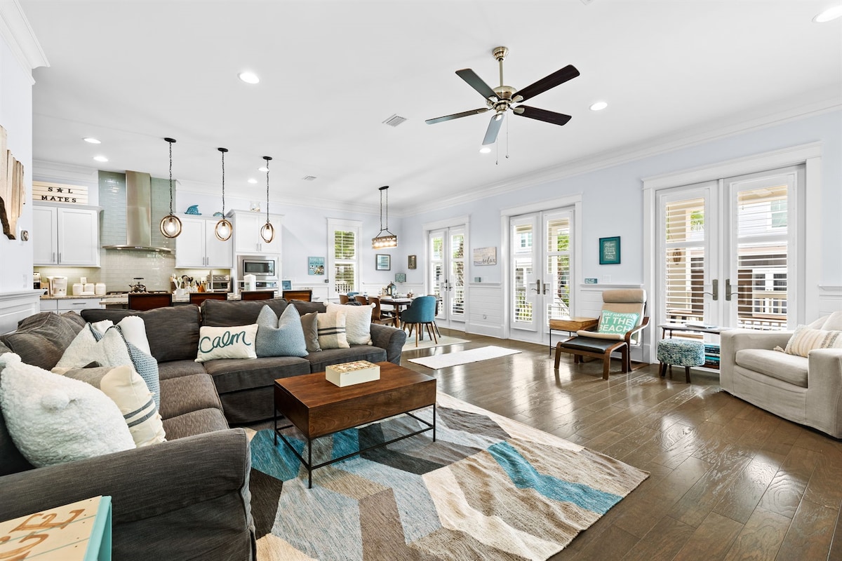 A spacious living area features a large sectional couch adorned with throw pillows and a coffee table at the center. Light wood flooring complements the soft blue walls. A ceiling fan hangs overhead, while natural light enters through double doors leading to the outdoor space.