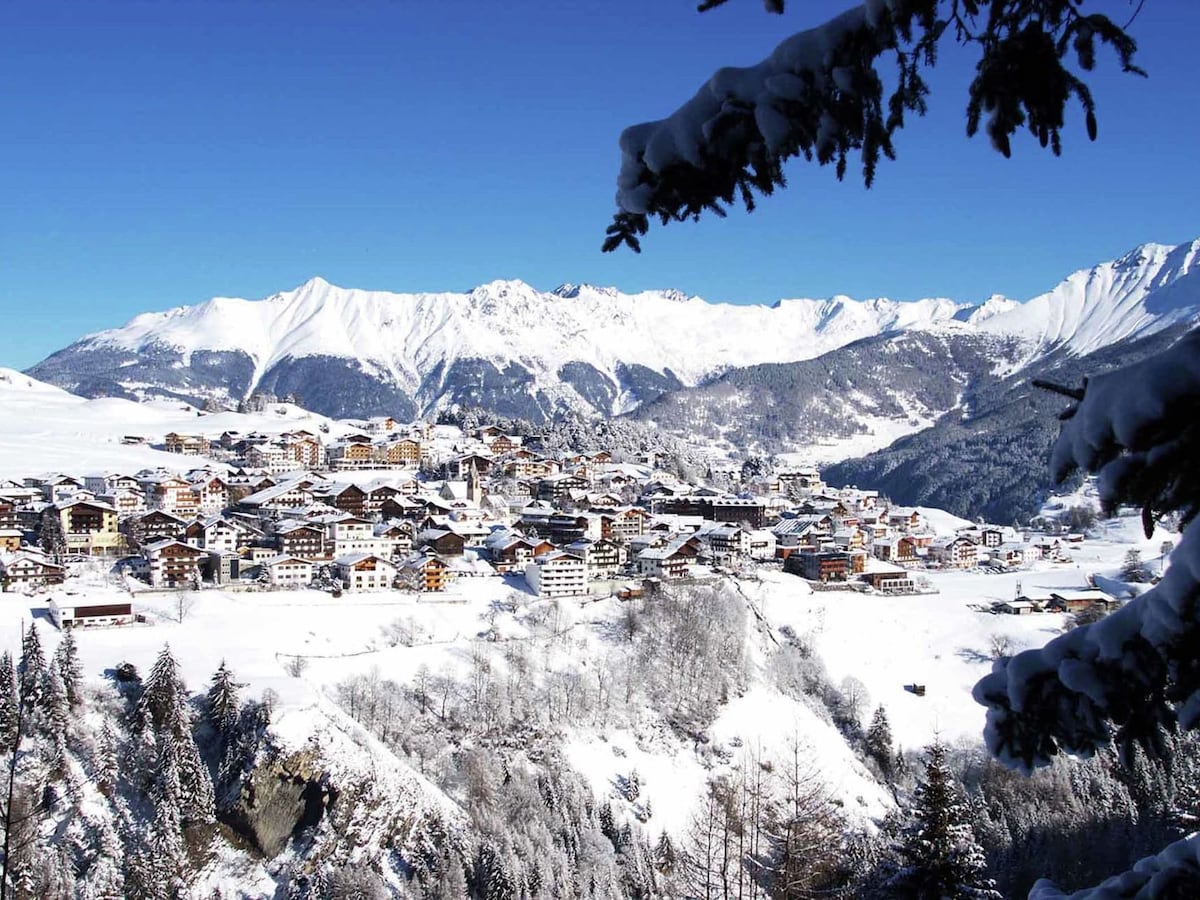A scenic view features a charming village nestled among snow-covered mountains. The roofs of colorful buildings peek through a blanket of white snow, while majestic peaks rise in the background against a clear blue sky.