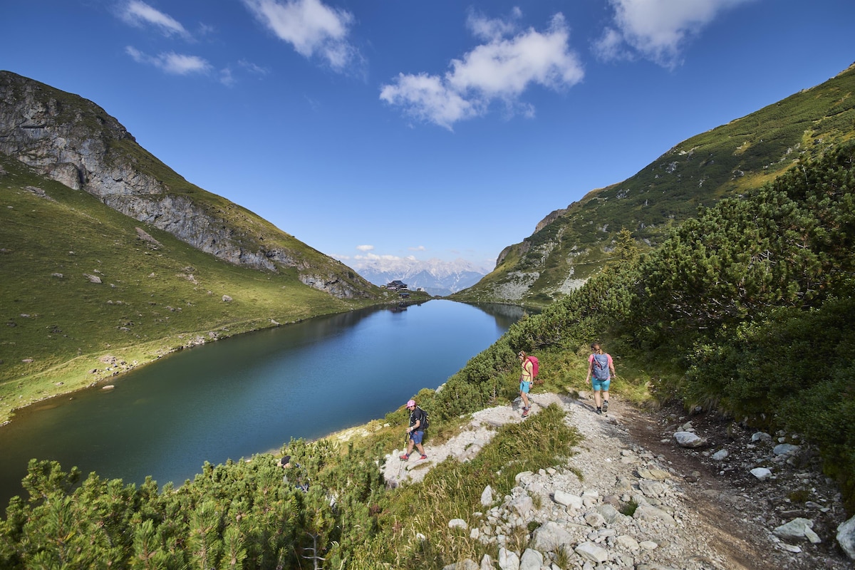 A serene lake is surrounded by lush green mountains under a clear blue sky. A well-defined walking path leads along the water's edge, where individuals are seen exploring the scenic landscape. The vibrant colors of the vegetation contrast harmoniously with the tranquil lake surface.