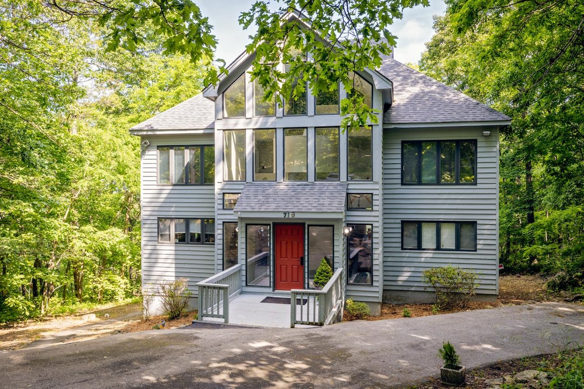 A three-story home is nestled among lush greenery, featuring a large front porch and a striking red door. The facade showcases numerous large windows that allow natural light to fill the interior. The driveway leads to a spacious entrance, emphasizing accessibility.