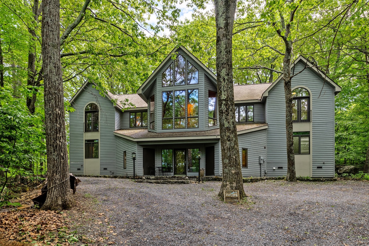 The exterior of a spacious home is highlighted by large glass windows that reflect the surrounding greenery. It is situated in a wooded area, with a gravel driveway leading to the entrance. A sense of privacy is conveyed by the tall trees flanking the property.