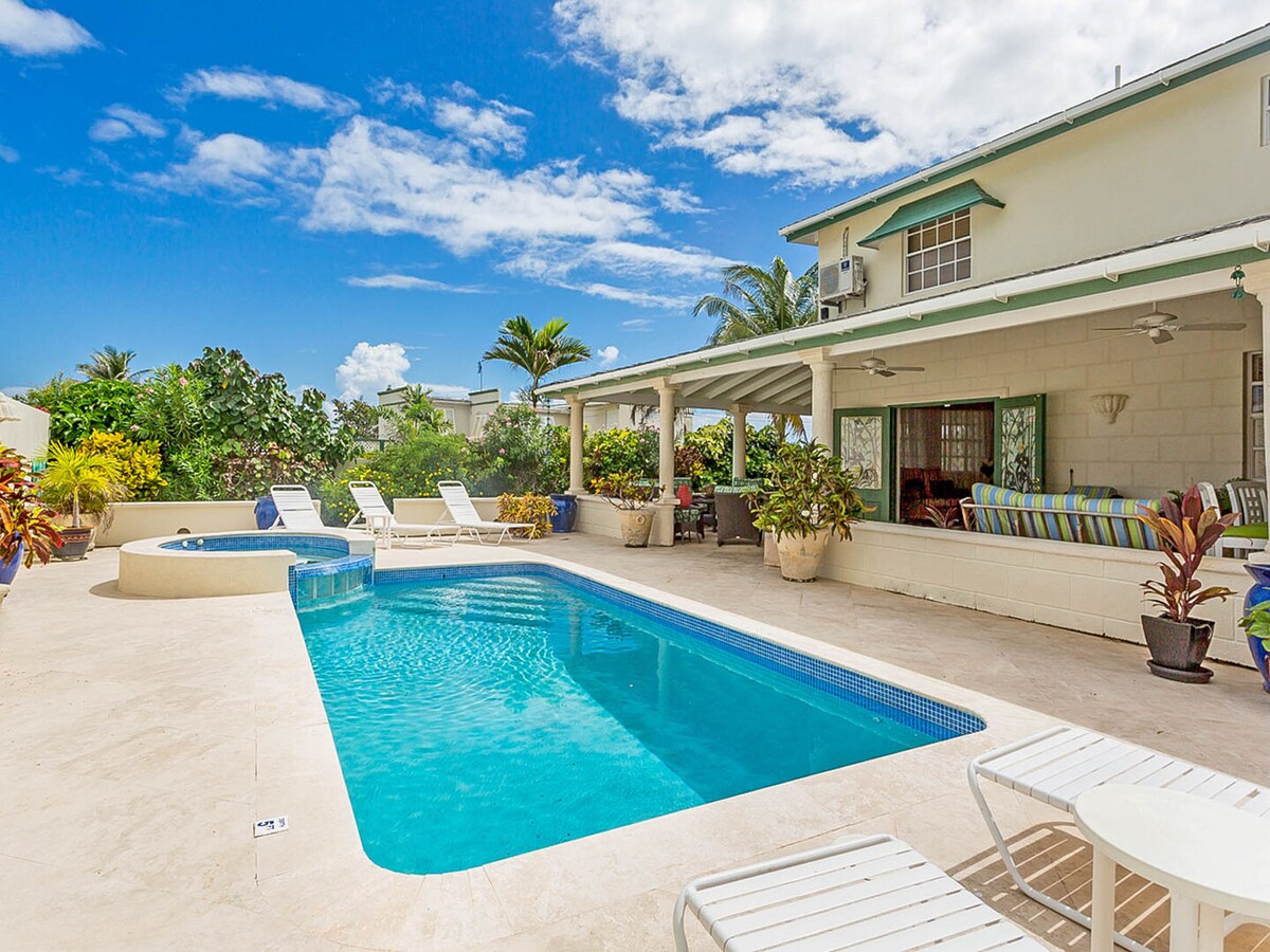 An expansive pool deck features a large swimming pool surrounded by lounge chairs and tropical plants. A separate hot tub offers added relaxation. The covered patio provides shaded seating with views of palm trees and a bright blue sky.