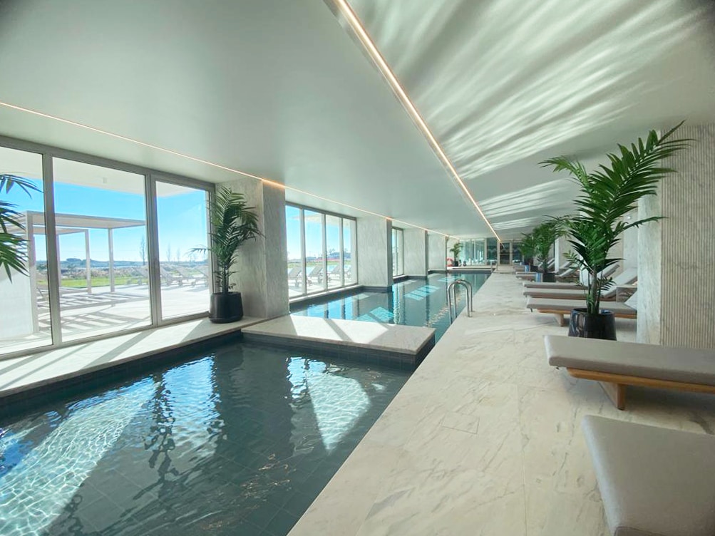 An indoor pool area features a sleek design, with large glass windows allowing natural light to flow in. Lush plants are strategically placed beside the pool, while comfortable lounge chairs line the sunlit space, encouraging relaxation and tranquility.