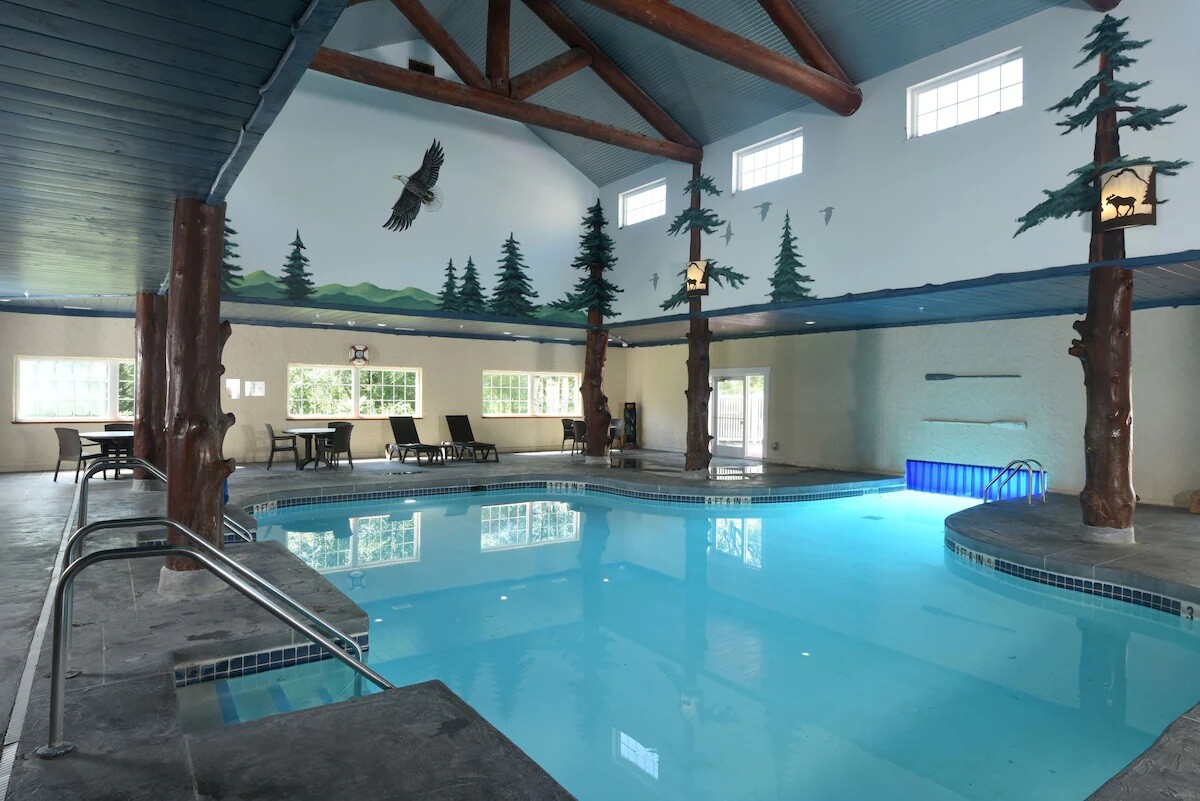 An indoor pool area is featured, with polished waters reflecting the ceiling's wood beams. Murals of trees and wildlife enhance the serene atmosphere. Surrounding lounge chairs provide space for relaxation, while large windows allow natural light to fill the area.