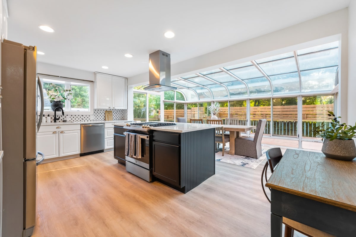 A modern kitchen is featured with sleek cabinetry, stainless steel appliances, and a large island with seating. Natural light fills the space through expansive windows, showcasing a dining area that connects to an outdoor patio. A touch of greenery adds a refreshing element.