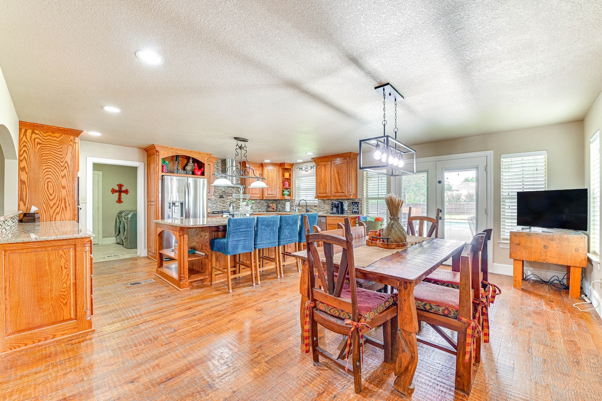 A spacious dining area features a large wooden table surrounded by ten padded chairs. The kitchen is open with wooden cabinetry, stainless appliances, and a large island with bar seating. Natural light floods the space through multiple windows, creating a bright atmosphere.