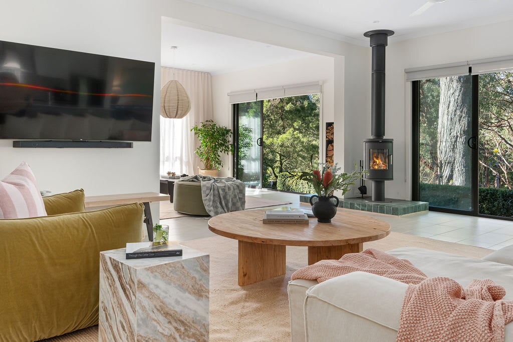 A spacious open living area is showcased, featuring a modern design with a cozy fireplace and large glass doors that connect to the outdoor scenery. A circular wooden coffee table sits in the center, complemented by a textured sofa and a decorative stone side table.