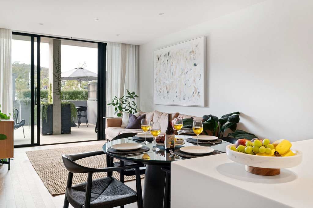 A modern dining area features a round table set for four, with beverages and fresh fruit displayed in a bowl. Large glass doors open to a terrace, allowing natural light to fill the space. A cozy seating area is visible in the background.