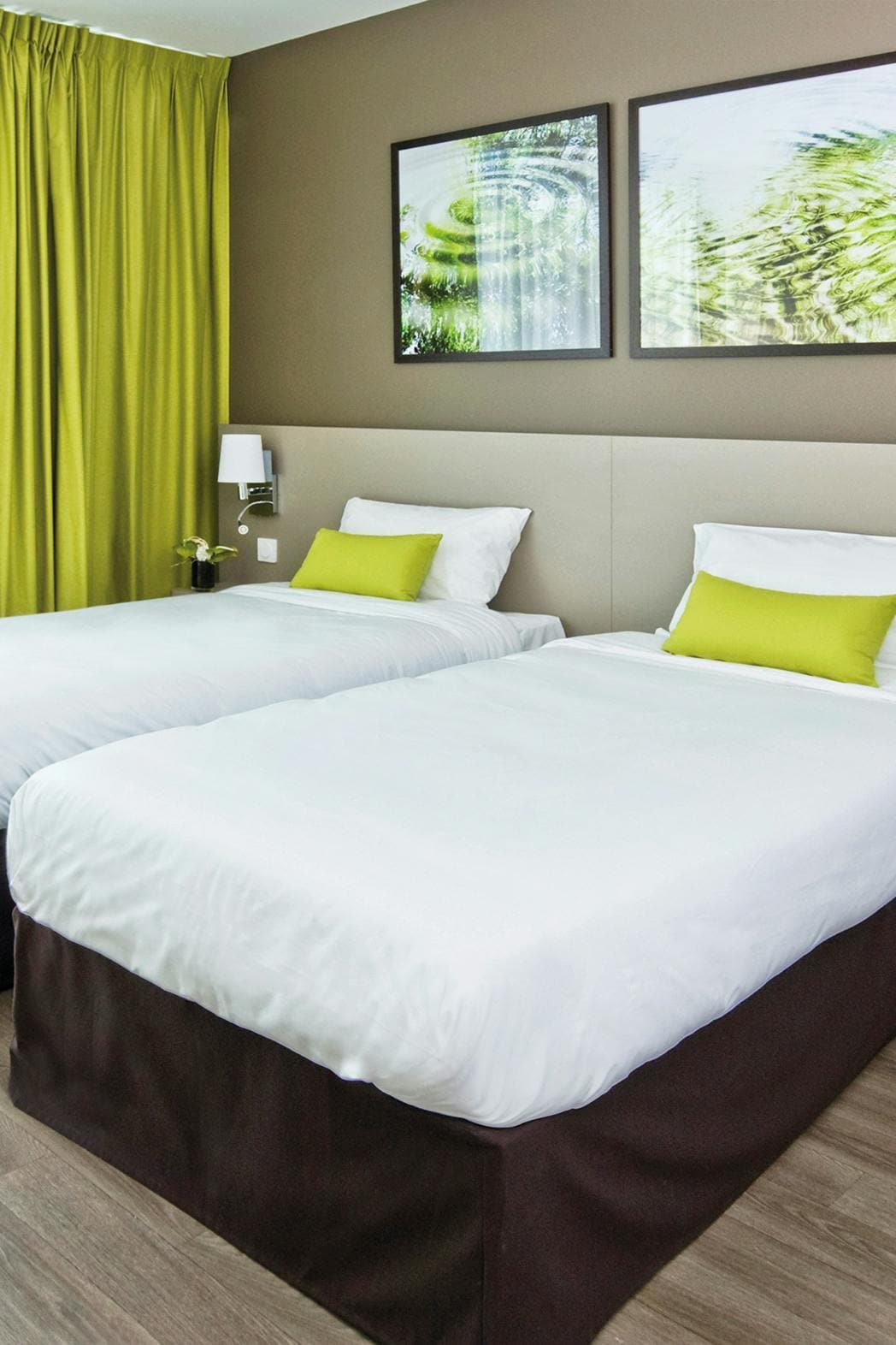 A well-lit room features two single beds dressed in crisp white linens, accented by bright green cushions. Floor-to-ceiling curtains in a matching hue frame the window. Two framed prints of greenery enhance the serene atmosphere.