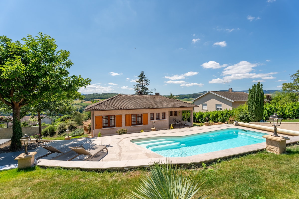 A spacious gîte is set against a backdrop of rolling hills, featuring a clear swimming pool surrounded by lounge chairs. Lush greenery and trees frame the property, while a well-maintained garden enhances the outdoor space. Soft clouds drift across a blue sky.