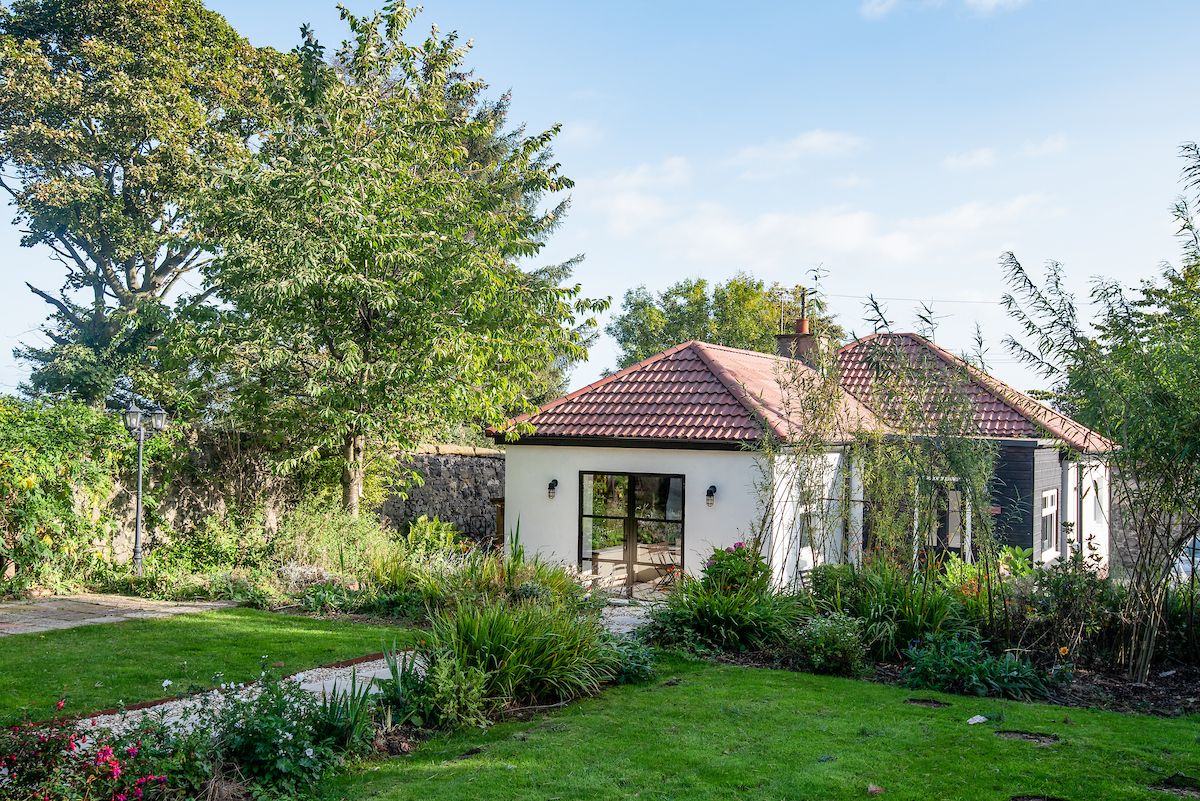 A single-story cottage with a red tiled roof is framed by lush green foliage and colorful plants. A pathway leads towards the entrance, showcasing a blend of natural elements and structured landscaping. The property has large windows that invite natural light into the interior.