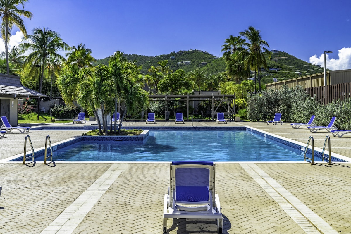 A large communal pool is framed by palm trees and lounge chairs, with a tropical landscape and rolling hills visible in the background. The pool water reflects the blue sky, and access steps are located on one side.