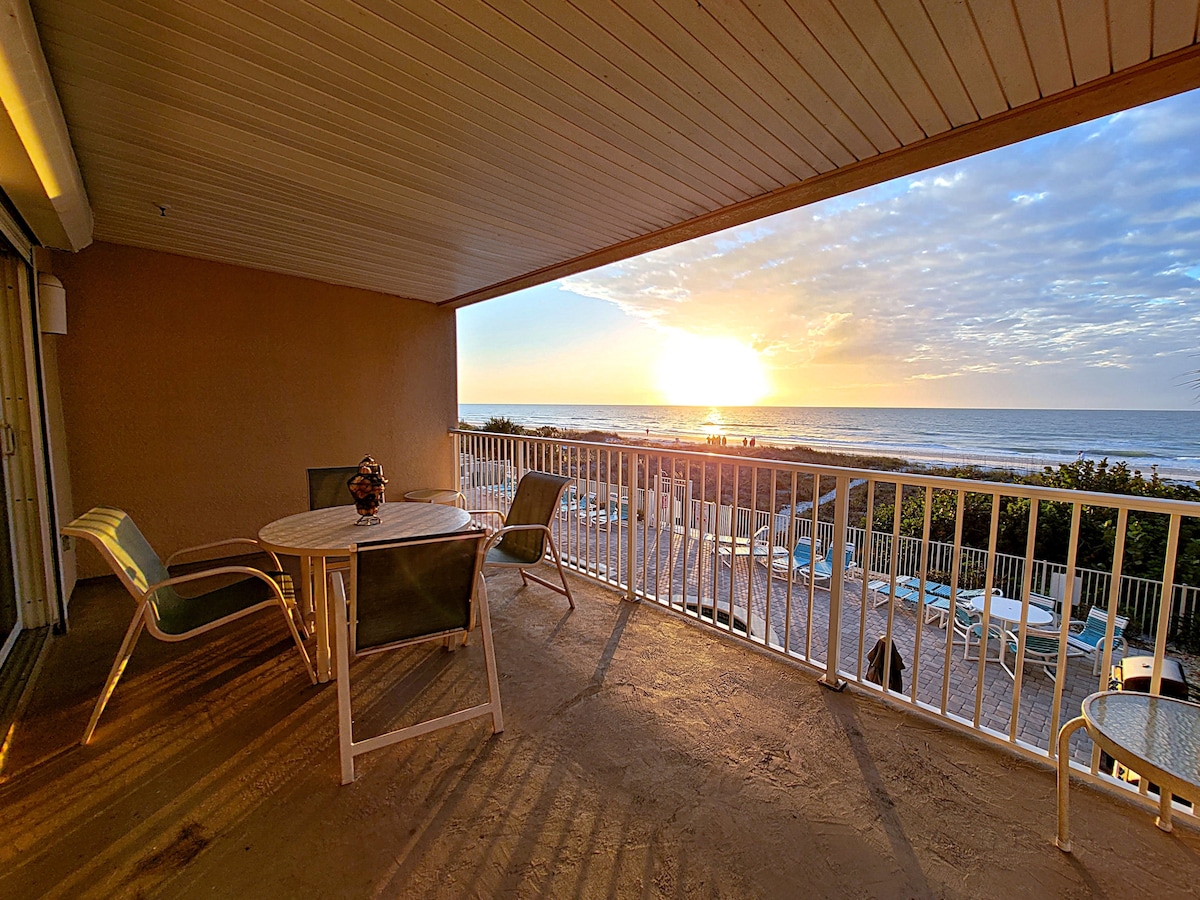 A spacious balcony offers a view of the beach and ocean, with a table and chairs positioned for enjoying the sunset. The horizon is illuminated with warm tones as the sun sets, while the nearby pool area is partially visible.