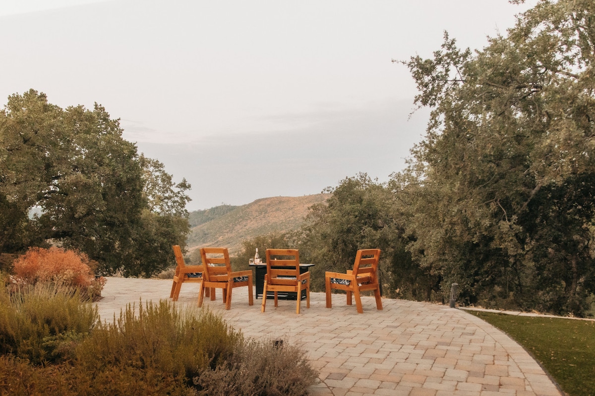 A spacious outdoor patio features four wooden chairs arranged around a central fire pit, offering an ideal setting for relaxation. Lush greenery and distant hills create a serene backdrop, while the stone pavers provide a solid surface for gatherings under the open sky.