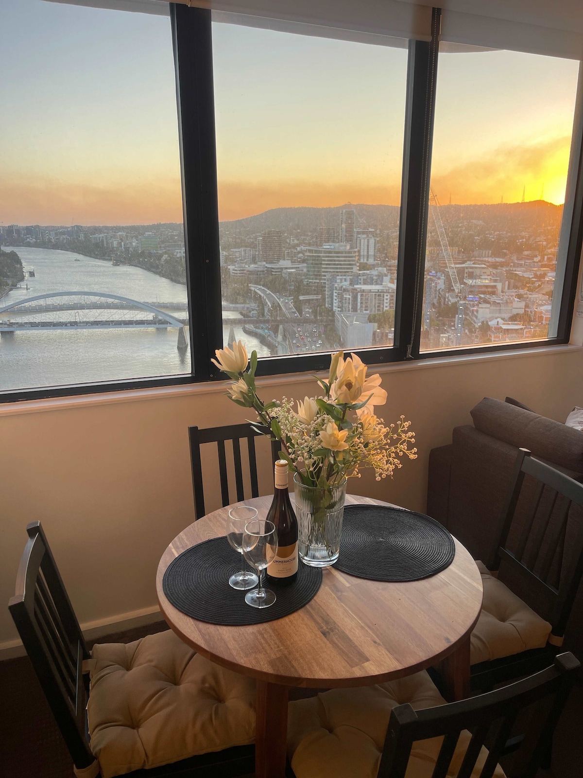 A small wooden dining table is set for two, featuring a vase with flowers and two glasses beside a bottle of wine. Floor-to-ceiling windows offer expansive views of the Brisbane River and sunset, enhancing the atmosphere of the space.