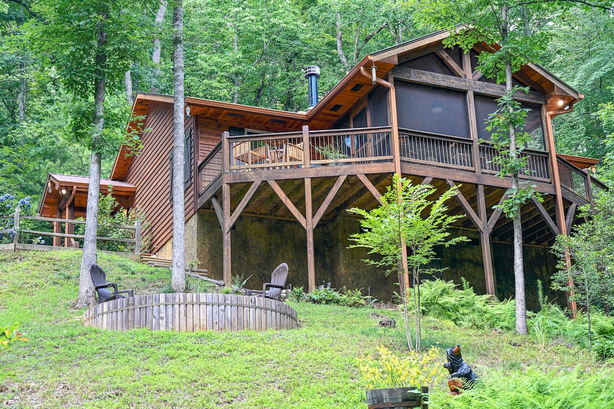 A wooden cabin is nestled among trees, featuring a spacious deck with a screened-in area and natural wood accents. A circular outdoor fire-pit with seating is visible nearby, surrounded by lush greenery, creating a serene and inviting setting.