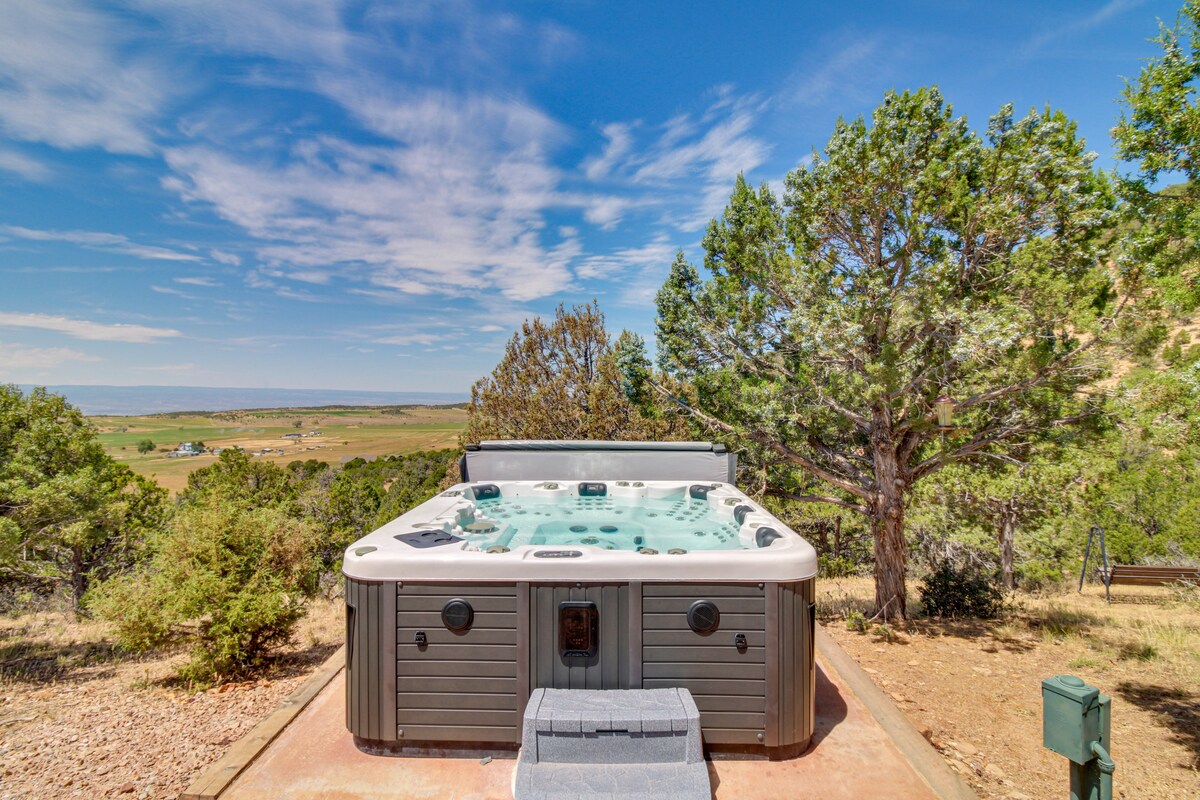 A hot tub is positioned on a concrete pad, surrounded by trees and hillside views. The setting features a clear sky, emphasizing a tranquil outdoor area suitable for relaxation.