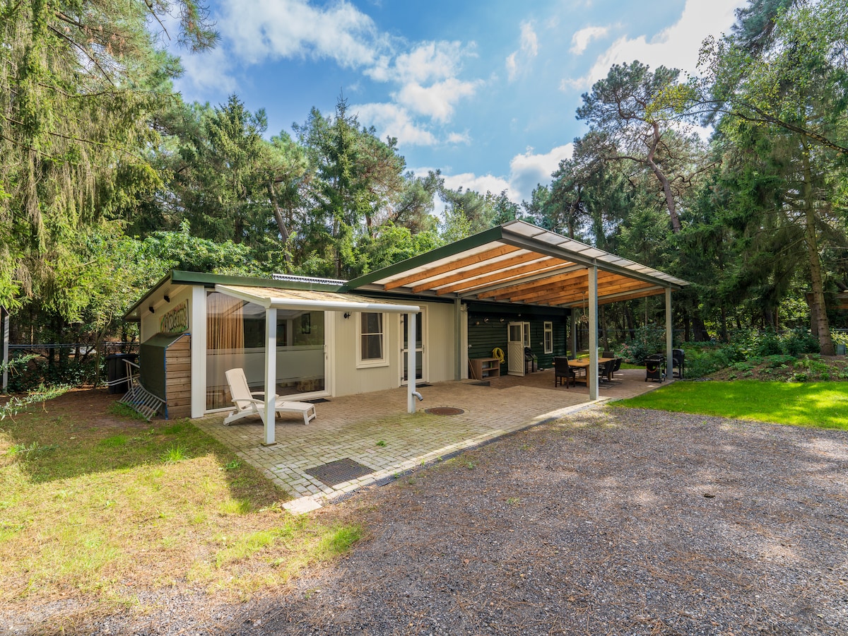 The holiday home features a covered terrace that extends to the outdoors, providing shaded seating areas. The entrance is framed by lush greenery, and a gravel path leads to the space. Large windows invite natural light into the inviting façade.