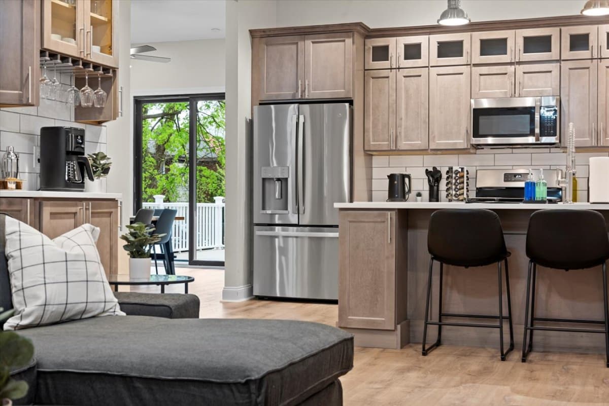 The kitchen features modern stainless steel appliances, including a refrigerator and microwave, along with wooden cabinetry. A large island with seating for two sets the stage for casual dining, while natural light floods the space through glass doors leading to a deck.
