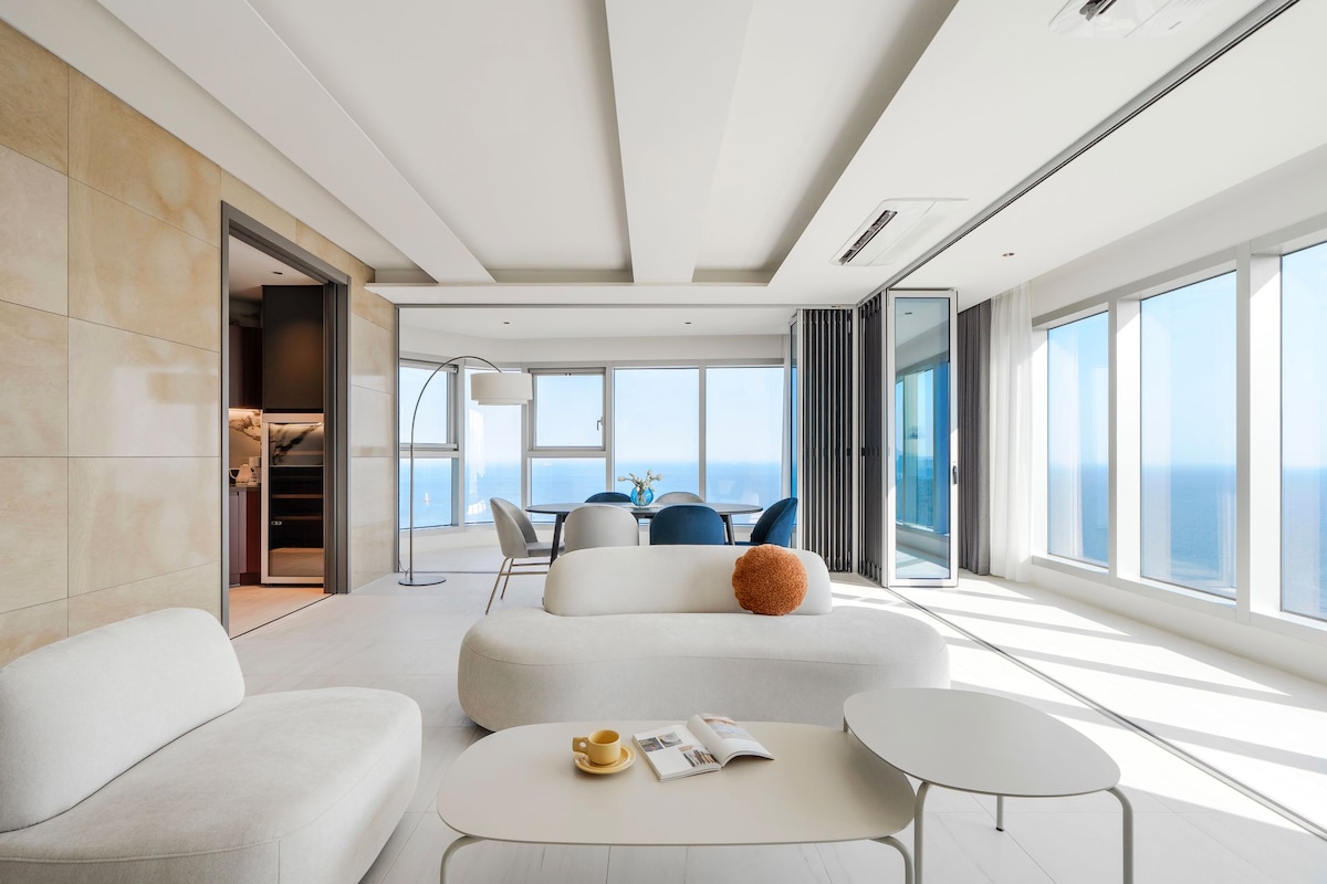 A spacious living area is highlighted by large windows offering expansive ocean views. Soft, contemporary furnishings include a white sofa and a round coffee table. Natural light enhances the ambiance, while a floor lamp adds a modern touch to the design.