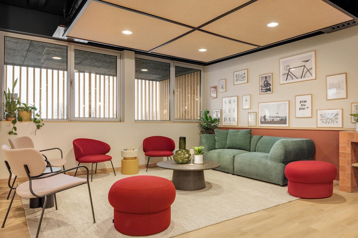 A welcoming lounge area is furnished with a green sectional sofa, two red ottomans, and several chairs in soft tones. Natural light filters through large windows adorned with horizontal blinds, while a round coffee table holds decorative items. Wall art adds character to the space.