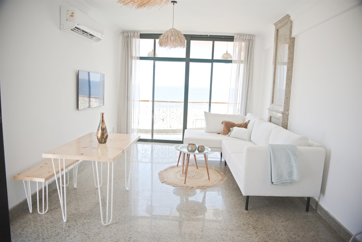 A modern living room features a comfortable white sofa, a light wood dining table, and decorative accents. Large windows frame the ocean view, allowing natural light to fill the space. A woven rug and minimalist decor enhance the room's fresh and airy ambiance.