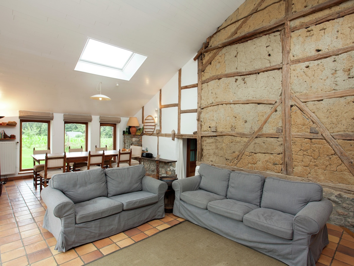 A spacious living area features two comfortable grey sofas and natural light enters through a skylight above. A dining table is visible in the background, complemented by rustic wooden beams that add character to the room. The floor is tiled with a warm, earthy hue.