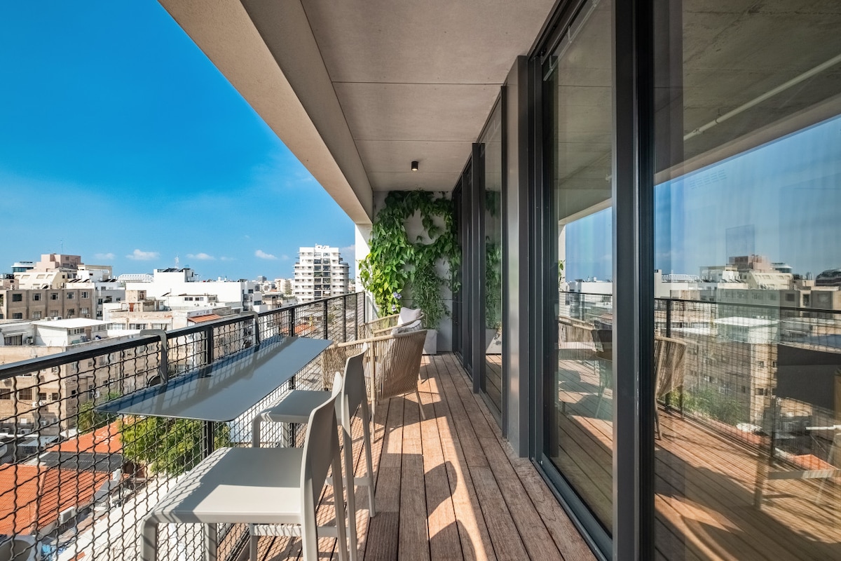 The private balcony features a spacious wooden deck complemented by a glass railing. A modern outdoor dining table with white chairs invites al fresco meals, while lush green plants add a touch of nature. Panoramic views of the city skyline are visible in the background.