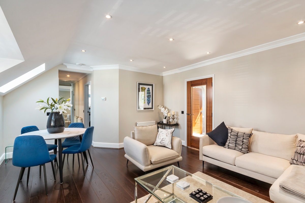A bright and open living area is featured, with a light color palette and hardwood flooring. A glass coffee table sits in the center, surrounded by a cozy sofa and two blue chairs. A dining table for four is positioned nearby, complemented by decorative wall art.