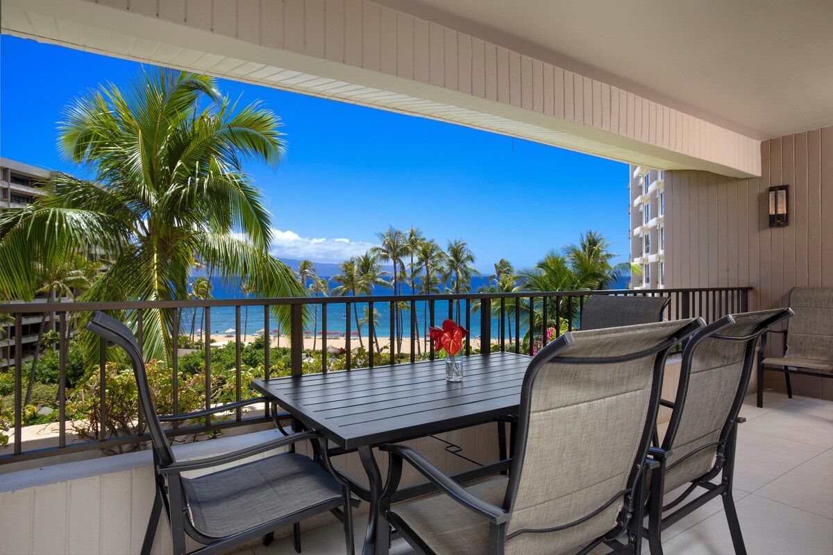 A spacious lanai offers a dining table with seating for four, overlooking lush palm trees and the ocean. Bright blue skies and the inviting shoreline are visible in the distance, creating a serene backdrop for outdoor gatherings.