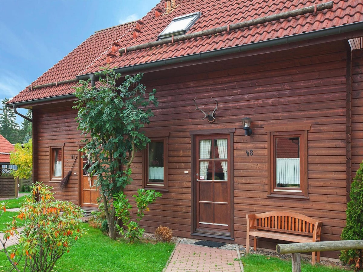 A charming log cabin with a brown wooden exterior is featured, highlighting a well-maintained entrance. A wooden bench sits invitingly on a brick walkway. Lush greenery surrounds the cabin, complemented by colorful shrubs and windows adorned with white curtains.