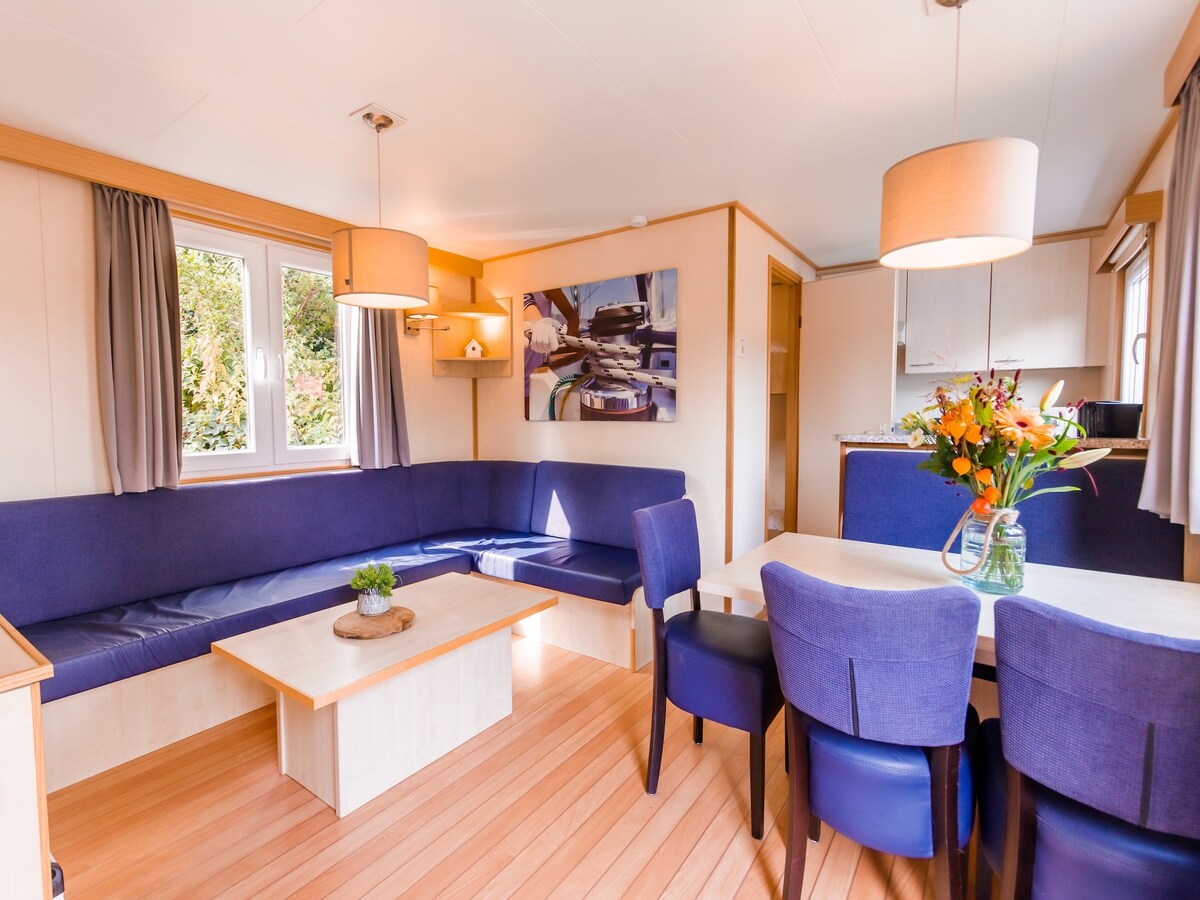 A modern living area features a spacious corner seating arrangement in deep blue upholstery, complemented by a dining table with four chairs. Light fixtures hang above, while a colorful artwork adds a decorative element to the wall. Windows allow natural light to fill the room.