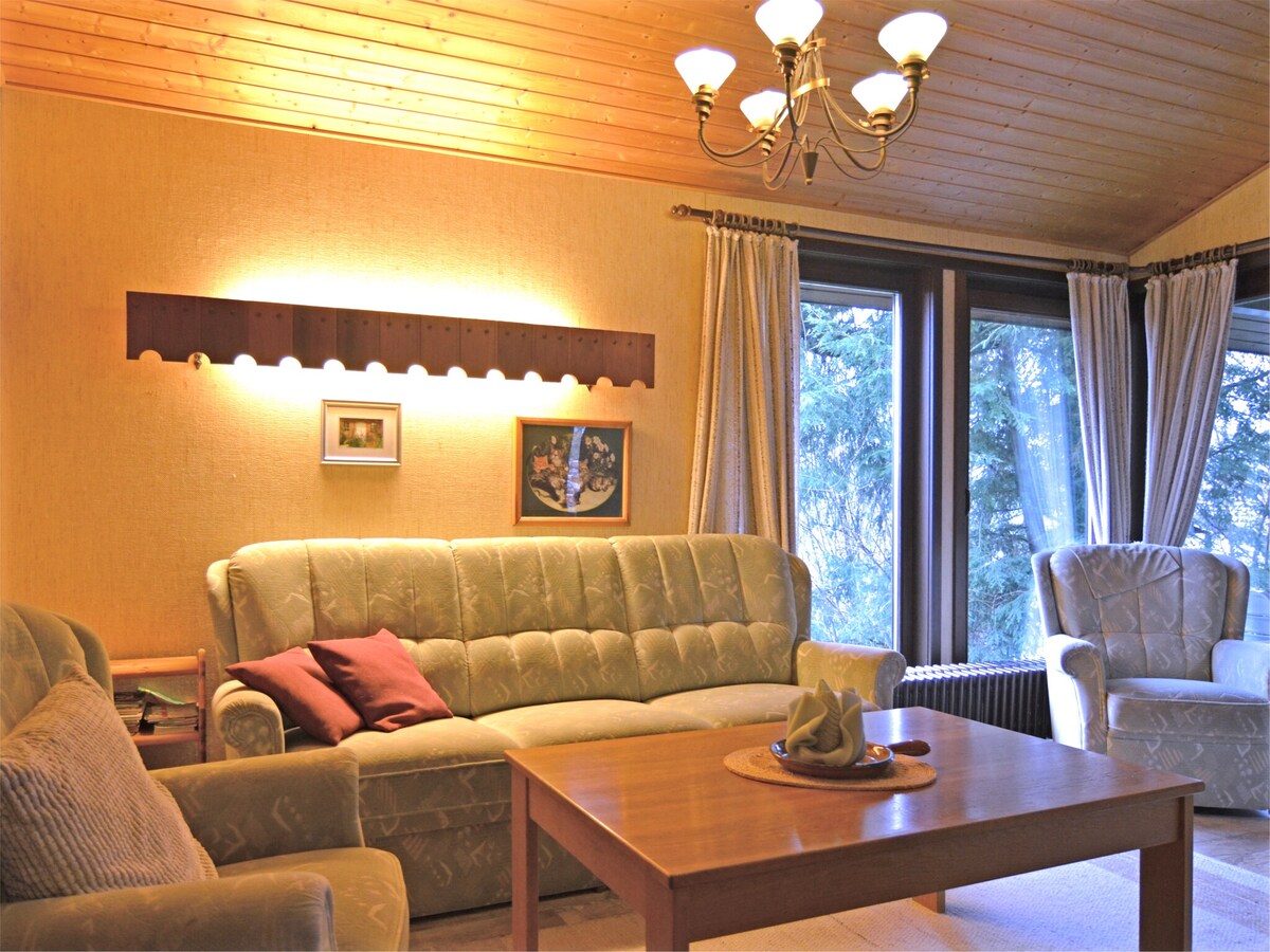 A comfortable living area is highlighted by a chandelier and warm lighting. A plush sofa and armchair offer seating, while a wooden table is centered for convenience. Large windows invite natural light and provide views of the surrounding greenery. Decorative wall art adds character.