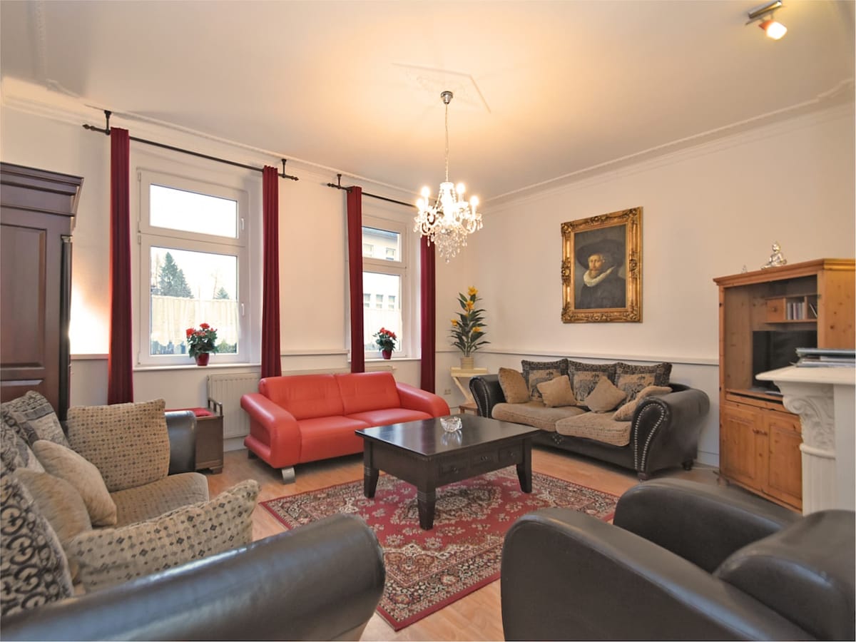 The living room showcases a mix of seating options, including a red sofa and dark leather armchairs arranged around a central coffee table. Natural light filters through large windows adorned with red curtains. A decorative chandelier hangs above, and a piece of wall art adds character to the space.