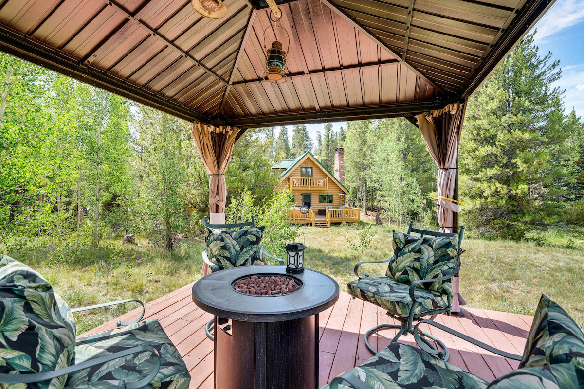 A covered gazebo is shown with comfortable seating and a central gas fire table surrounded by nature. The cabin can be seen in the background, framed by trees and grass that enhance the serene outdoor setting.