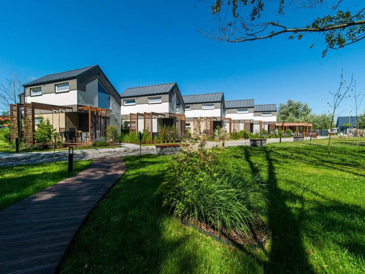 A row of modern holiday apartments is set against a clear blue sky. The structures feature large windows, surrounded by lush greenery and well-maintained gardens. A wooden path leads through the landscape, enhancing accessibility to the outdoor areas.
