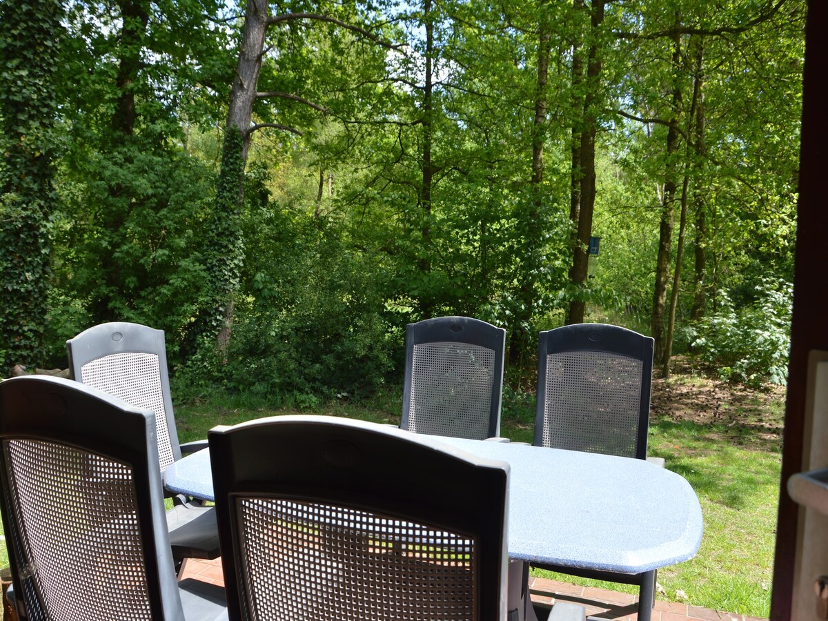 A dining set featuring a round table surrounded by six chairs is positioned on a patio. A lush backdrop of dense greenery and tall trees is visible, providing a natural setting for outdoor meals.