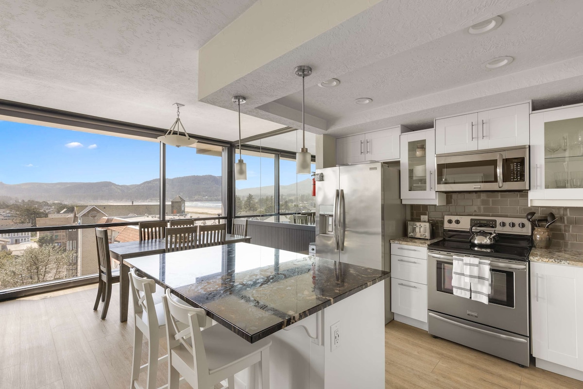 A modern kitchen features stainless steel appliances, granite countertops, and ample natural light from the large windows. A spacious island with seating for four is complemented by white cabinetry. Scenic views of the surrounding landscape are visible through the floor-to-ceiling windows.