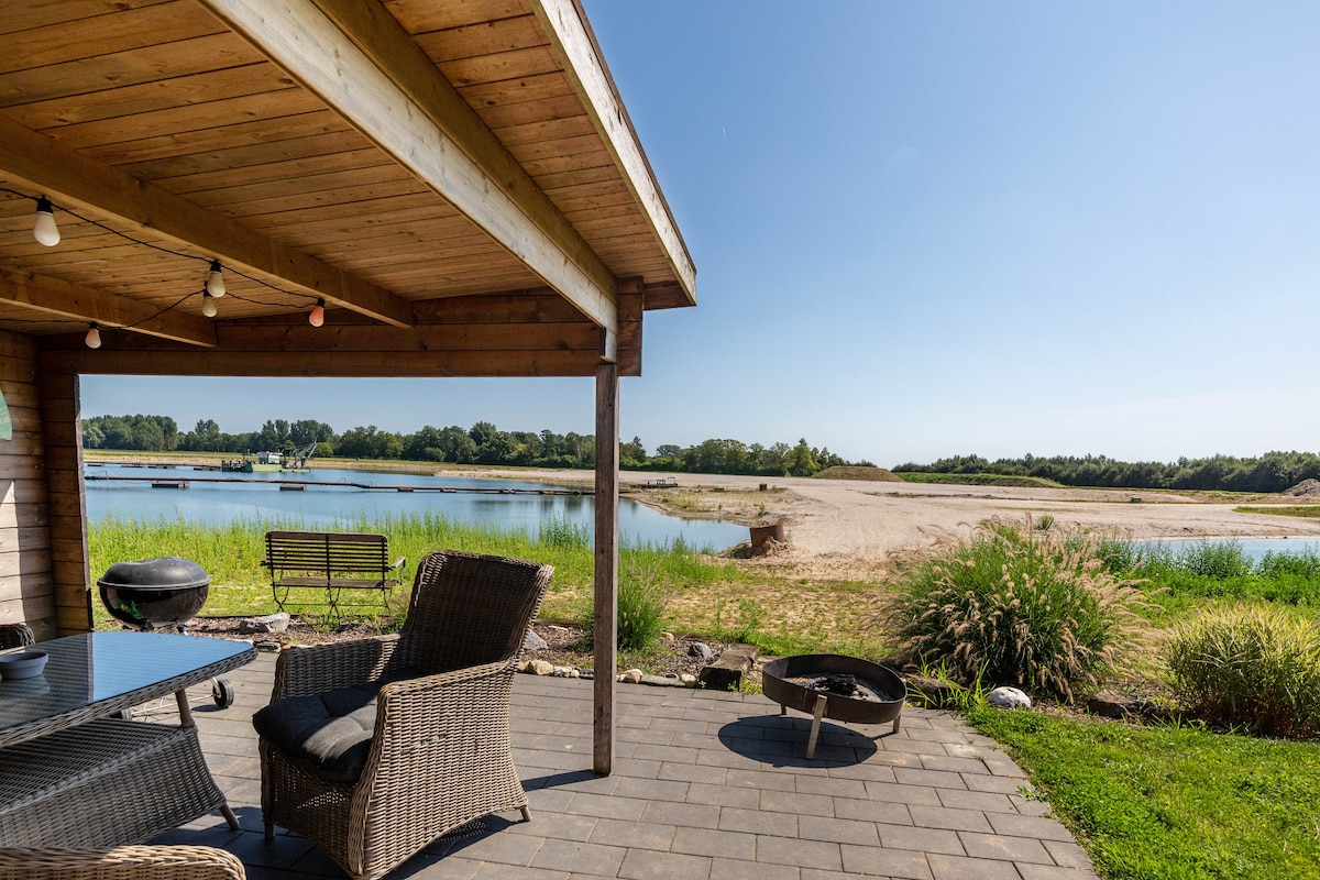 A spacious outdoor terrace features a wooden overhang with comfortable seating. A table is visible, alongside a fire pit. The view opens up to a serene lake and a grassy area, with a sandy spot in the background under a clear blue sky.
