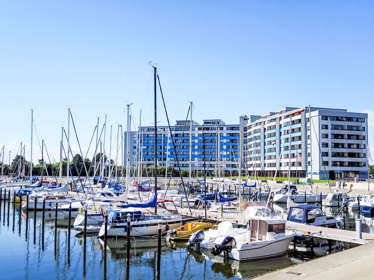 A marina is lined with various sailboats and motorboats, each secured at the dock. In the background, a modern multi-storey building features large windows and a blue façade, reflecting a clear blue sky.