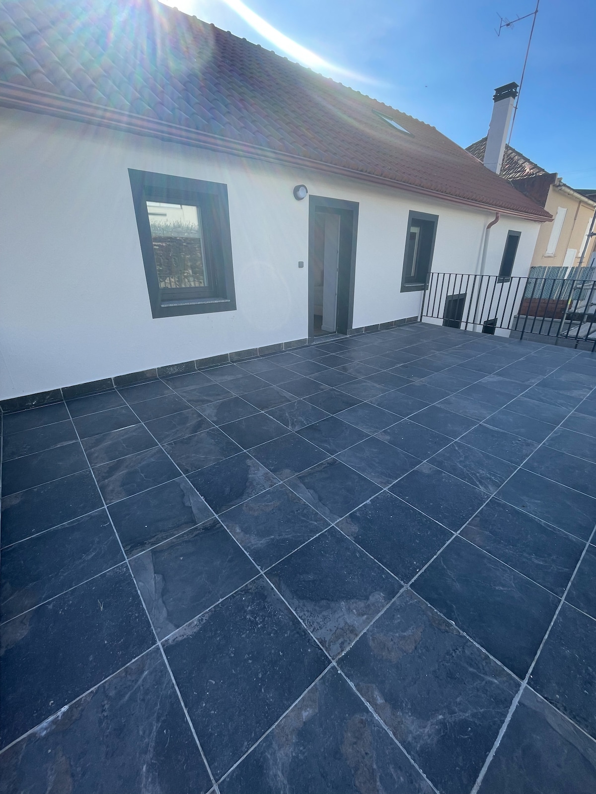 A spacious outdoor terrace is seen, featuring a dark tiled floor and low walls. Sunlight illuminates the area, which is bordered by a railing and includes large windows of the adjoining building in the background.