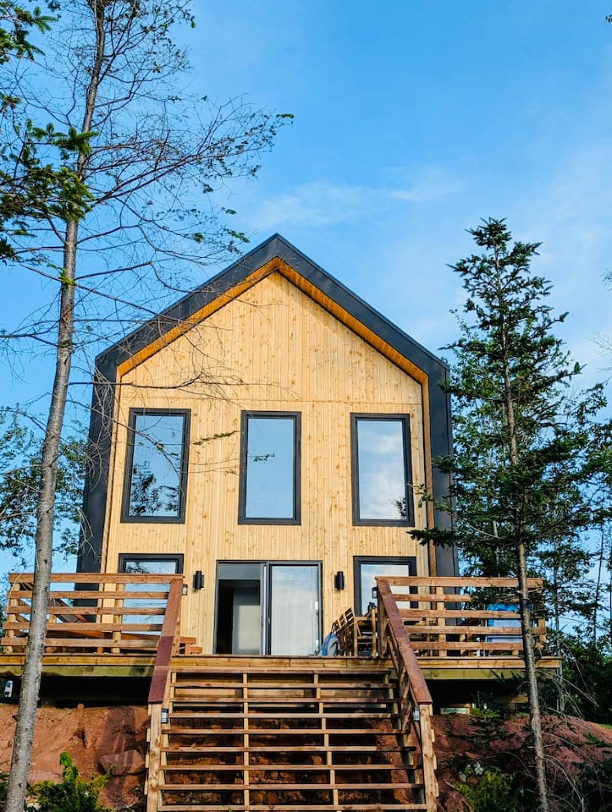 A modern chalet structure is showcased, featuring a pointed roof and large windows that reflect the natural surroundings. The exterior is clad in light wood, complemented by a wooden staircase leading to an elevated deck. Trees are visible around the property.